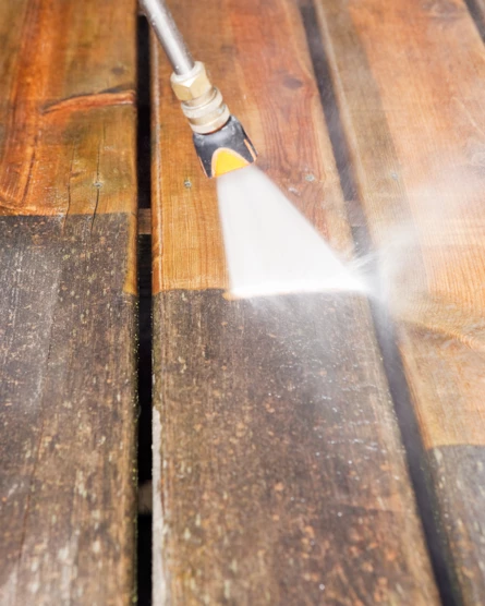 Houten terras wordt gereinigd met een hogedrukreiniger; groot contrast tussen gereinigd en vuil oppervlak.